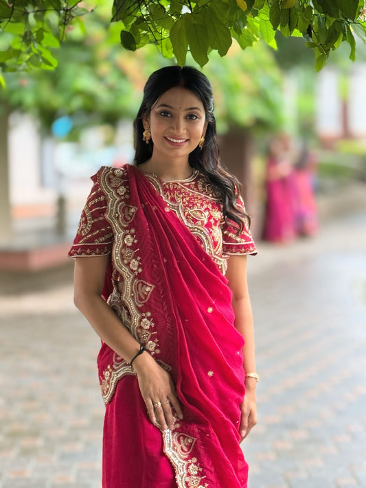 Woman in a red saree with gold hand embroidery standing outdoors under greenery.